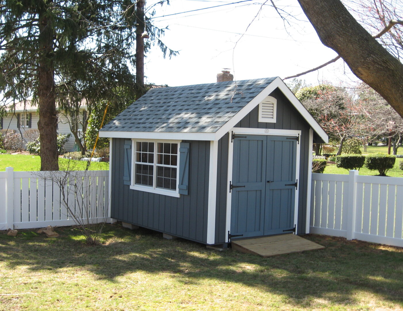 Classic Cottage shed with blue siding, white trim, white windows with blue shutters, blue double doors, a ramp, and light gray asphalt roofing along a white fence in a backyard.