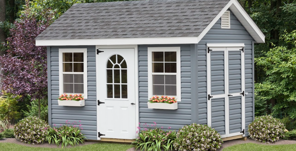 a frame shed with blue siding, white windows and doors, flower boxes, and shrubs along the perimeter