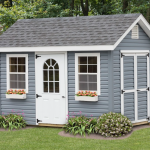 a frame shed with blue siding, white windows and doors, flower boxes, and shrubs along the perimeter